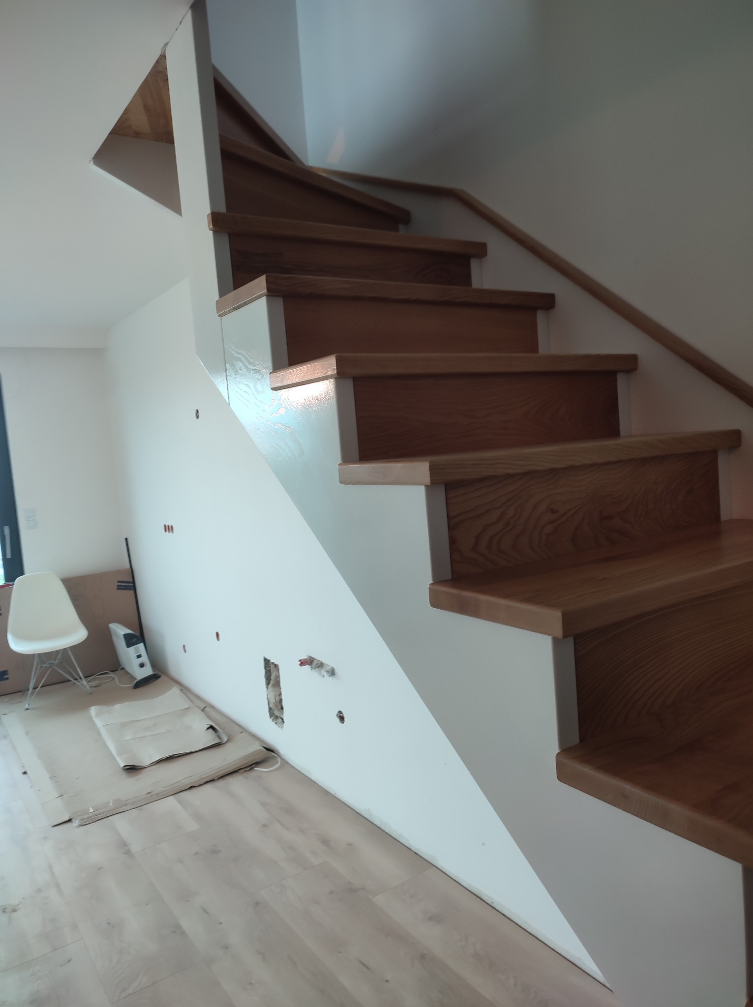 Drewniane schody z białymi podstopnicami podczas montażu, widoczne przygotowania do instalacji elektrycznej w ścianie, zabezpieczona podłoga i krzesło w tle.