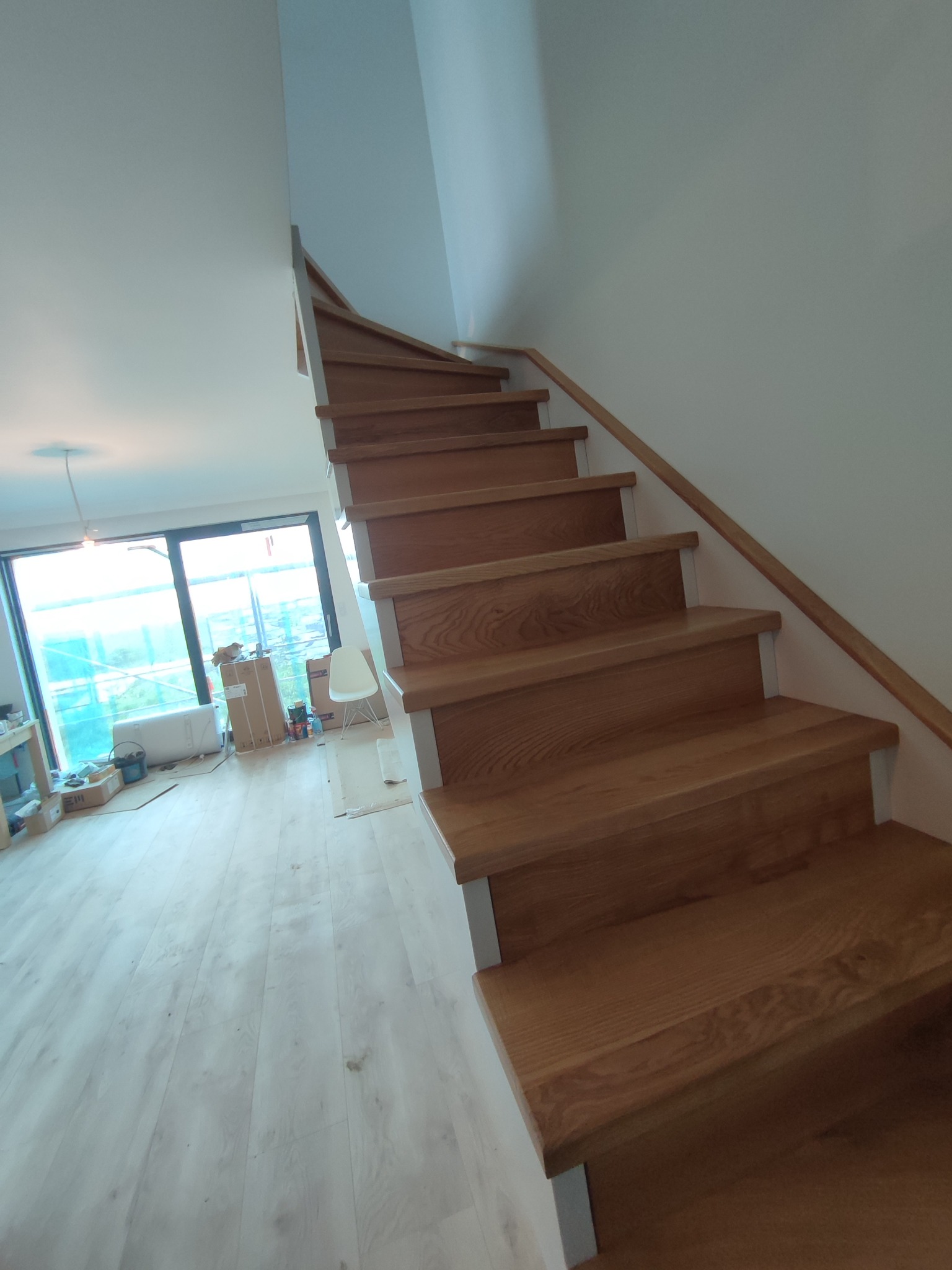 Drewniane schody z jasnymi podstopnicami i poręczą, widok z dołu, w tle okno z widokiem na budowę.