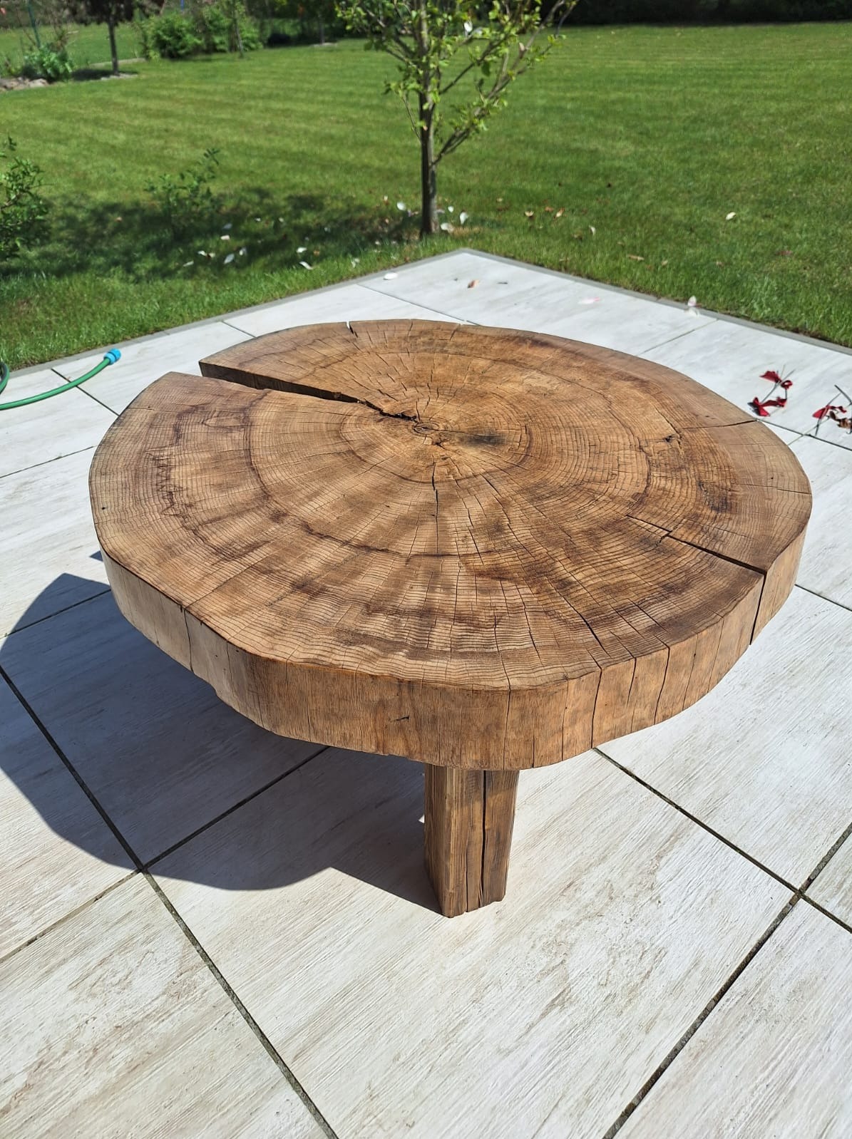 Stolik kawowy z grubego pnia drzewa z widocznymi słojami, umieszczony na tarasie z jasnych płytek, w tle trawnik i drzewo.