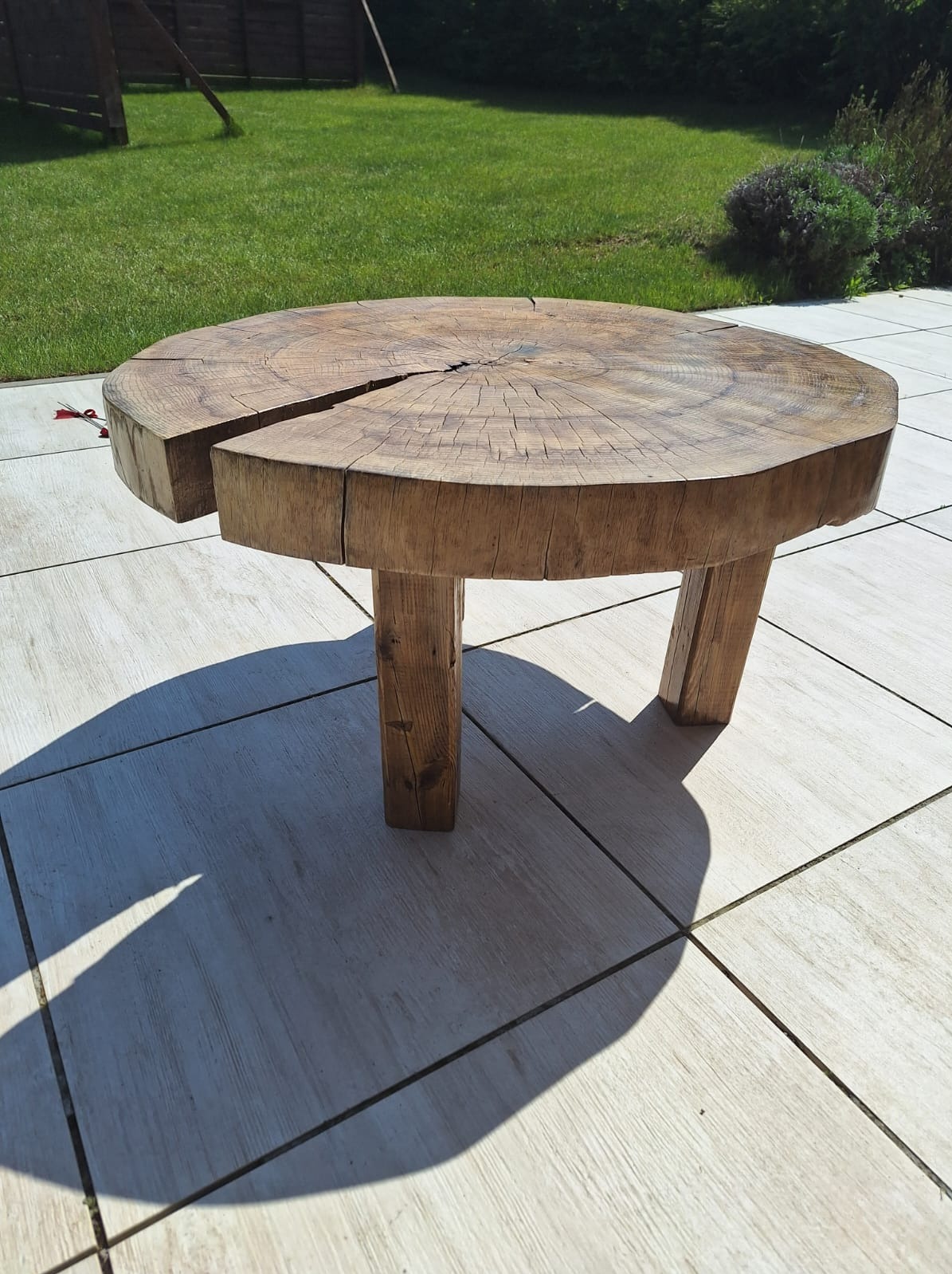 Stolik kawowy z pnia drzewa z wyraźnym pęknięciem na blacie, trzy proste, kwadratowe nogi, ustawiony na jasnej, kwadratowej terakocie w ogrodzie z trawnikiem w tle.