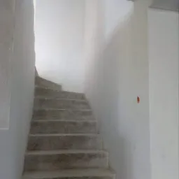 Surowe betonowe schody w trakcie budowy, prowadzące w górę w jasnym, minimalistycznym wnętrzu, z widocznymi niedoskonałościami na ścianach i schodach.