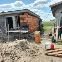 Zakład ogólnobudowlany - Budowa: Murowane ściany z pustaków ceramicznych na zaprawie, widoczny fragment budynku w trakcie wznoszenia, piasek, betoniarka i narzędzia budowlane.