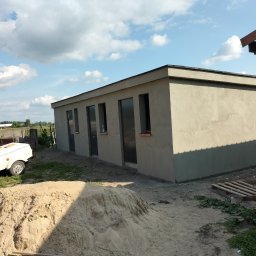 Zakład ogólnobudowlany - Nowo otynkowany budynek gospodarczy z czterema oknami w szarej elewacji, sterta piasku i przyczepka na pierwszym planie, błękitne niebo z chmurami w tle.