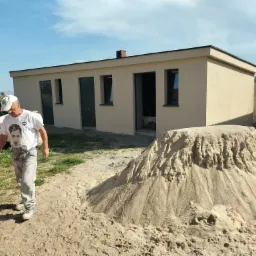 Parterowy budynek z beżową elewacją, widoczny stos piasku i pracownik w tle. Budynek z oknami i drzwiami, na tle zieleni i błękitnego nieba. Ukończony etap prac.