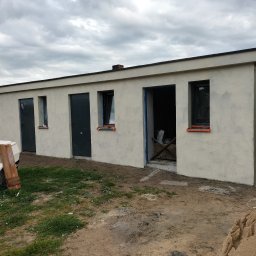 Zakład ogólnobudowlany - Niski budynek po tynkowaniu elewacji z trzema drzwiami i oknami, przy jednym wejściu widoczne narzędzia, na trawie przyczepka samochodowa.