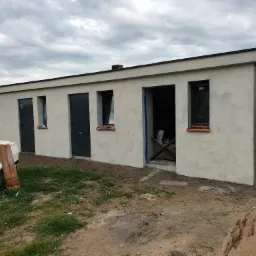 Niski budynek po tynkowaniu elewacji z trzema drzwiami i oknami, przy jednym wejściu widoczne narzędzia, na trawie przyczepka samochodowa.