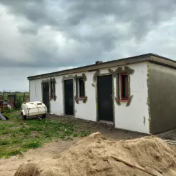 Budynek w trakcie ocieplania: białe płyty styropianowe na ścianach, częściowo pokryte szarą warstwą kleju, widoczne okna i drzwi. Na pierwszym planie hałda piasku.