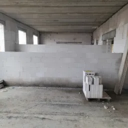 Wnętrze budynku w trakcie budowy. Widoczne ściany z białych bloczków, surowy betonowy strop, otwory okienne i paleta z bloczkami. Surowy stan deweloperski.