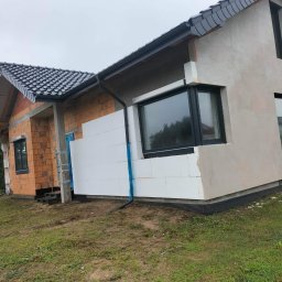 Zakład ogólnobudowlany - Dom w trakcie ocieplania: fragment ściany z cegły, fragment ocieplony styropianem, fragment otynkowany. Widoczne okna z czarnymi ramami i czarna rynna.