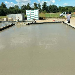 Zakład ogólnobudowlany - Wylewka fundamentowa w trakcie prac. Widoczny pracownik wyrównujący powierzchnię betonu, bloczki H+H, teren budowy i częściowo zaparkowany samochód dostawczy.