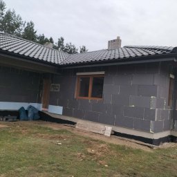 Zakład ogólnobudowlany - Budowa domu jednorodzinnego w trakcie ocieplania. Widoczne szare bloczki styropianowe na ścianach, okna z drewnianymi ramami i dach pokryty szarą dachówką.