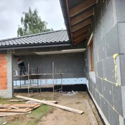 Zakład ogólnobudowlany - Budowa domu z szarego bloczka: widok na ścianę z oknem, częściowo ocieploną, z rusztowaniem i pracownikiem w tle. Widoczne deski i ceglany filar.