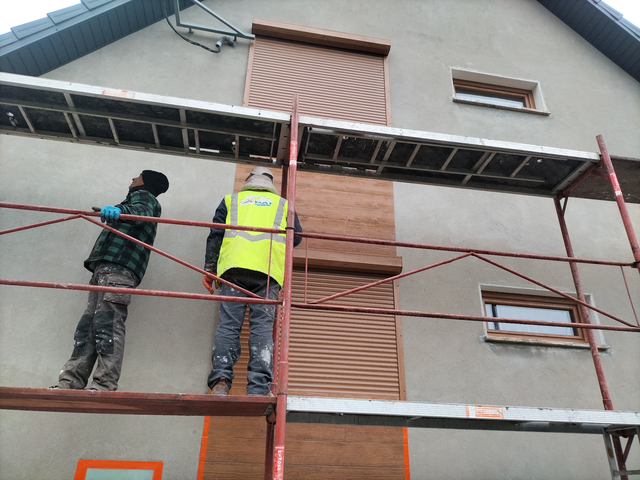 Pracownicy na rusztowaniu montują brązowe panele elewacyjne imitujące drewno na ścianie domu z szarą elewacją, widoczne okno i roleta.