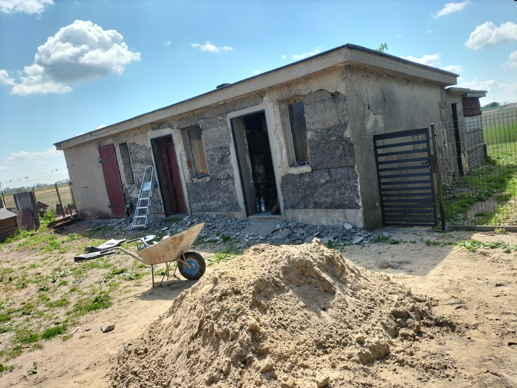 Niedokończony budynek z uszkodzoną elewacją, sterta piasku i taczka na pierwszym planie, drabina oparta o ścianę, częściowo odsłonięte mury.