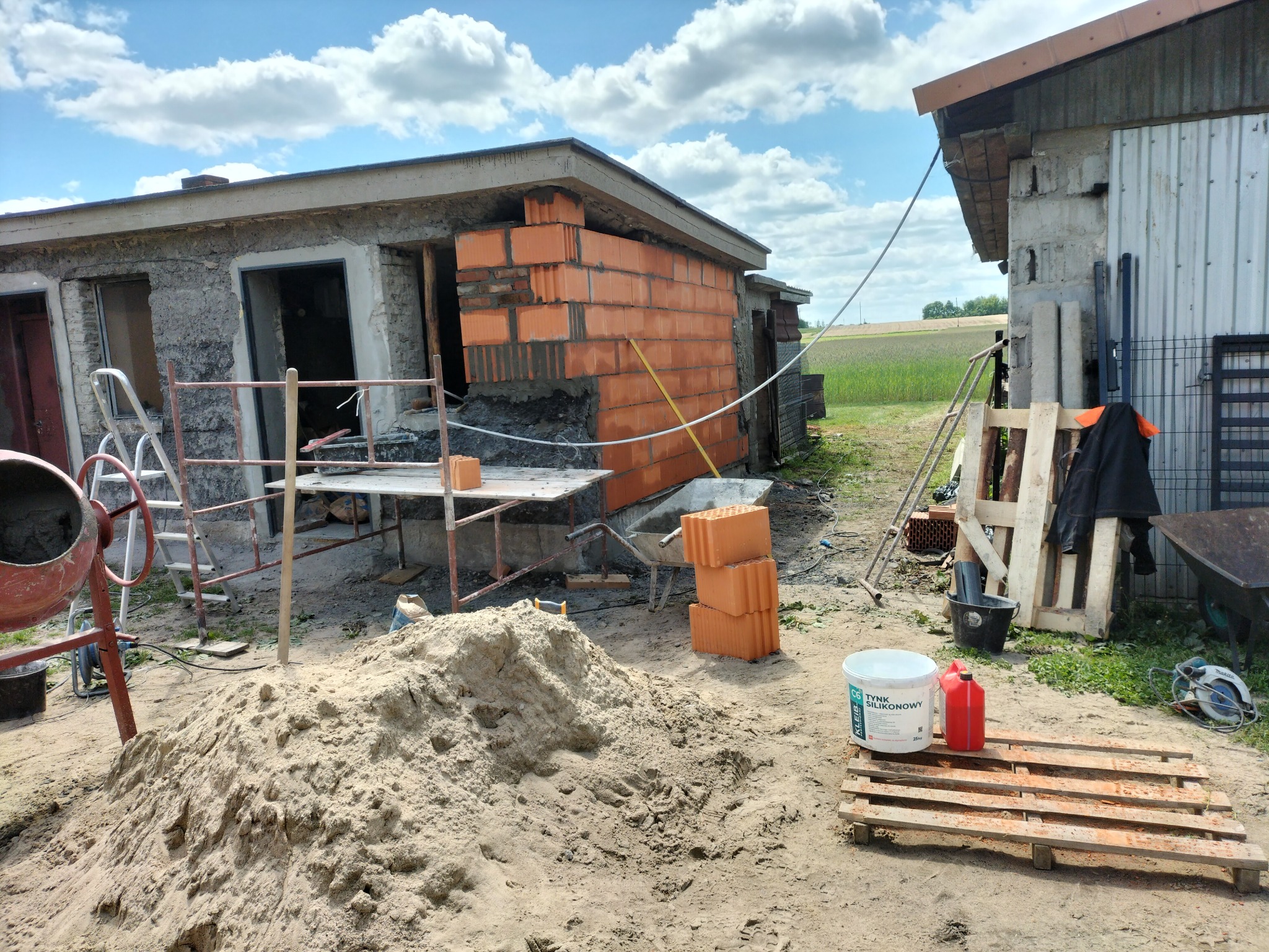 Budowa: Murowane ściany z pustaków ceramicznych na zaprawie, widoczny fragment budynku w trakcie wznoszenia, piasek, betoniarka i narzędzia budowlane.