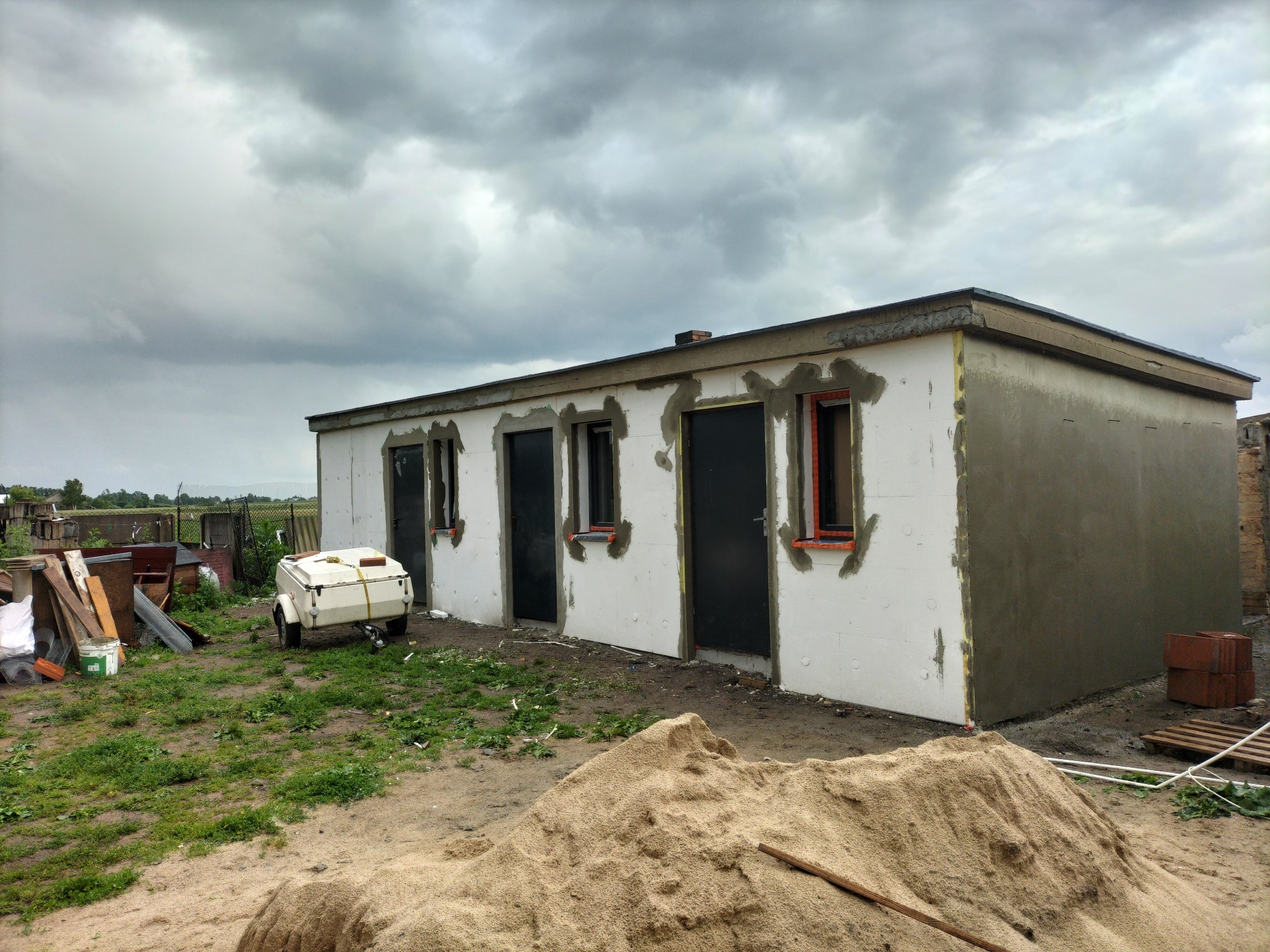 Budynek w trakcie ocieplania: ściany pokryte styropianem, fragment elewacji z szarą warstwą kleju. Na pierwszym planie sterta piasku pod pochmurnym niebem.