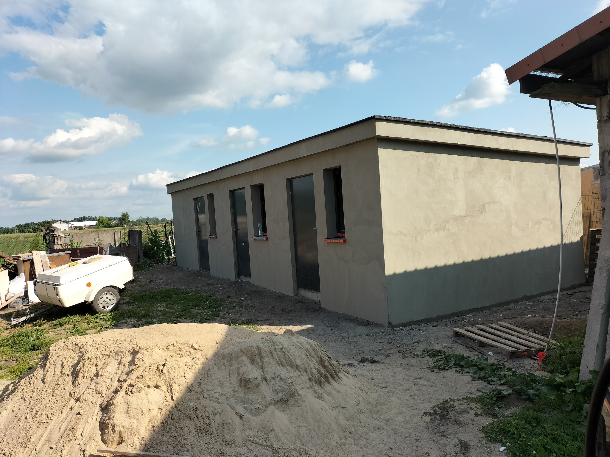 Nowo otynkowany budynek gospodarczy z czterema oknami w szarej elewacji, sterta piasku i przyczepka na pierwszym planie, błękitne niebo z chmurami w tle.