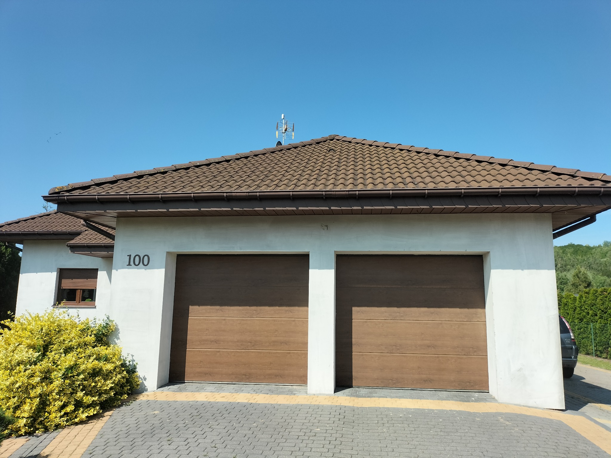 Nowoczesny dom z dwoma brązowymi bramami garażowymi, numerem 100 na białej elewacji i brązowym dachem pod błękitnym niebem. Żółty krzew przed budynkiem.