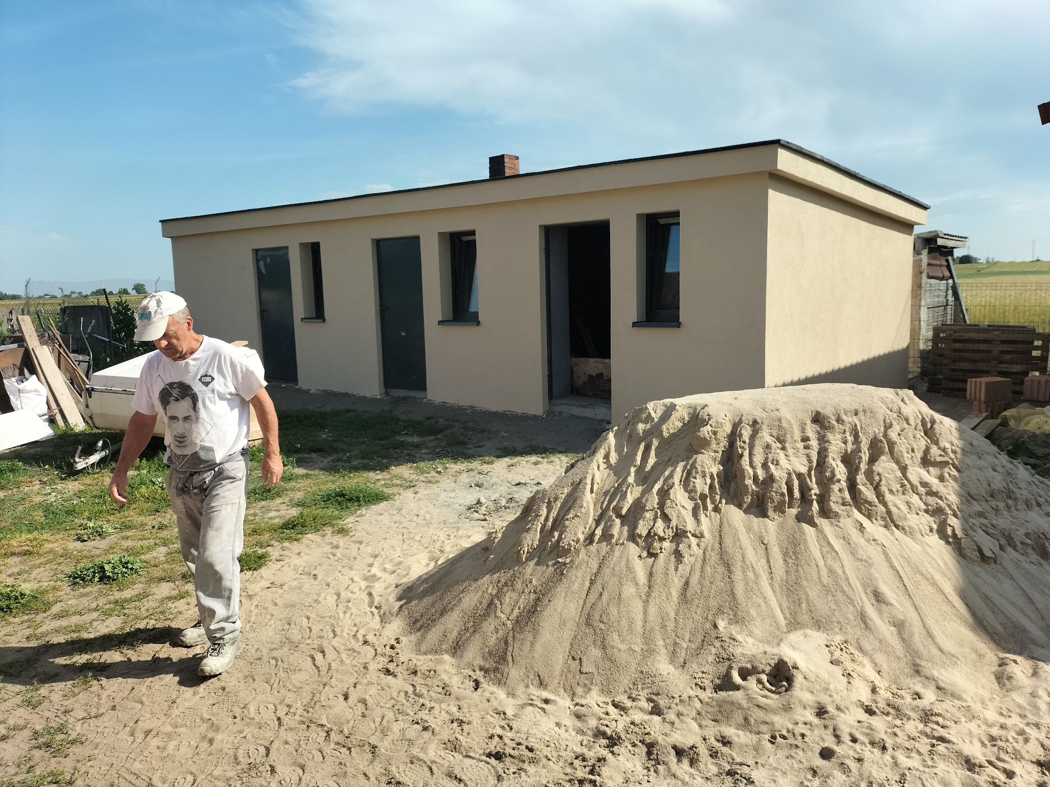 Parterowy budynek z beżową elewacją, widoczny stos piasku i pracownik w tle. Budynek z oknami i drzwiami, na tle zieleni i błękitnego nieba. Ukończony etap prac.