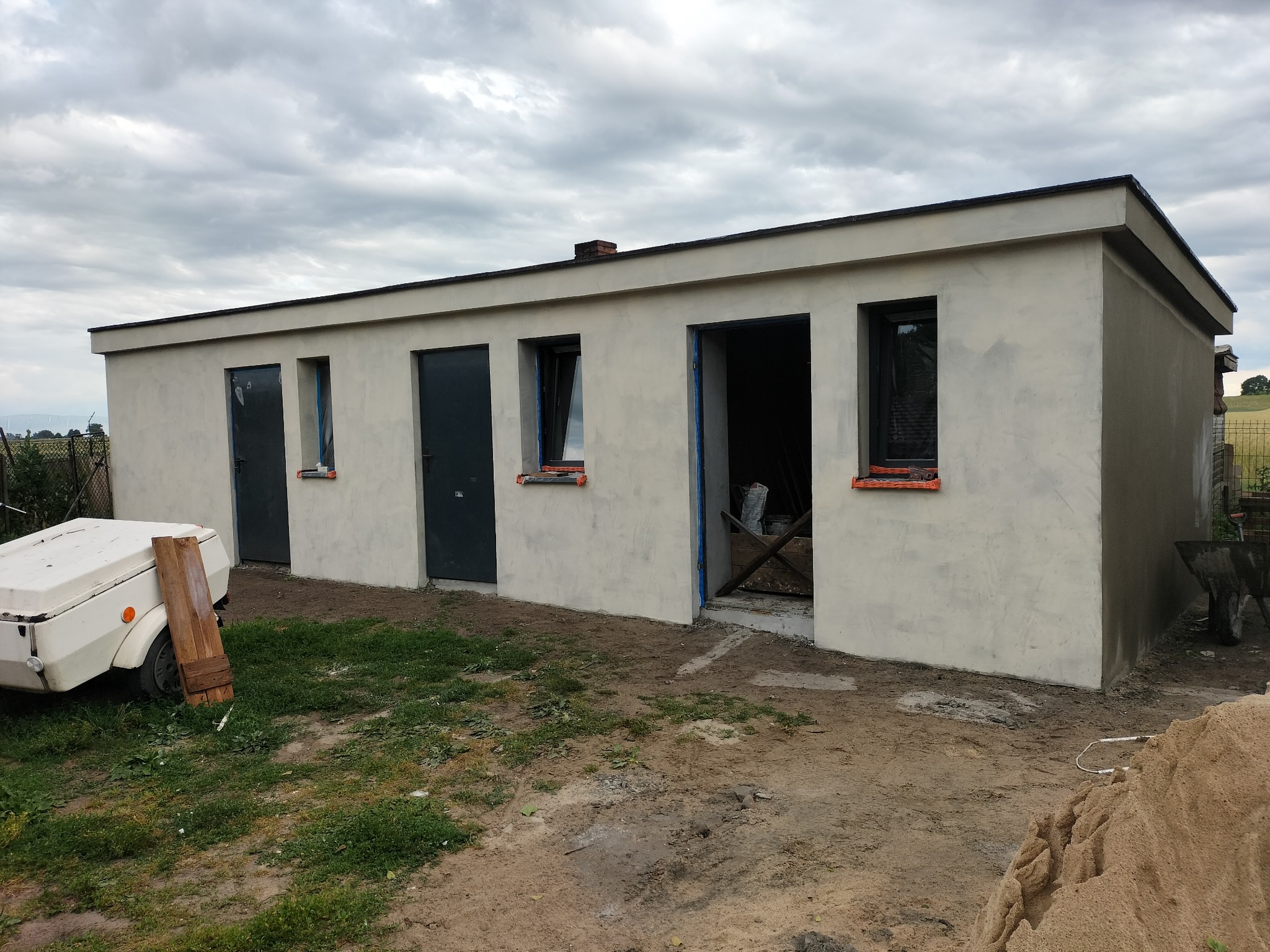 Niski budynek po tynkowaniu elewacji z trzema drzwiami i oknami, przy jednym wejściu widoczne narzędzia, na trawie przyczepka samochodowa.