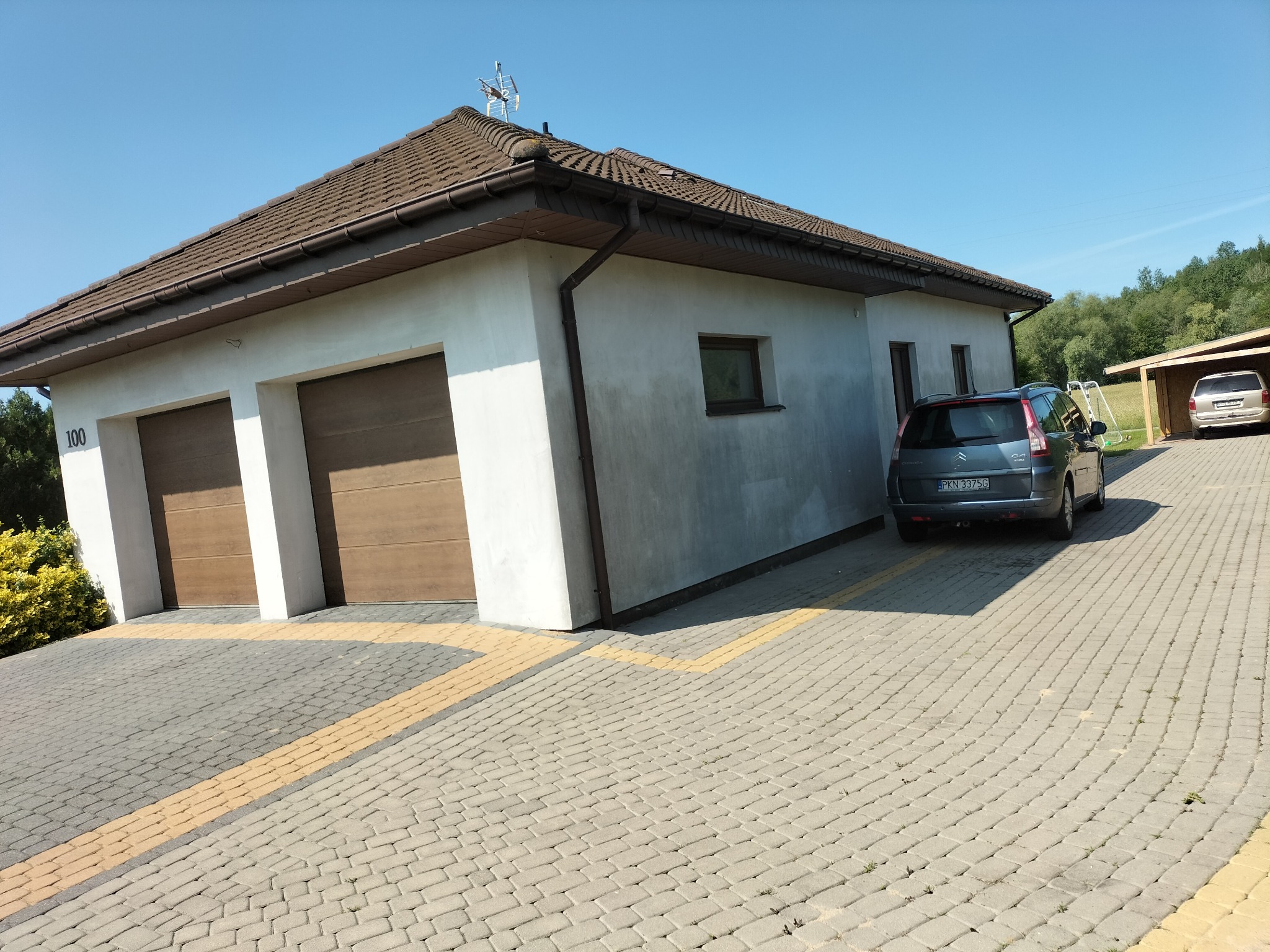 Parterowy dom z brązowym dachem i dwoma garażami, samochód zaparkowany obok. Widoczna kostka brukowa na podjeździe i zieleń w tle. Antena na dachu.