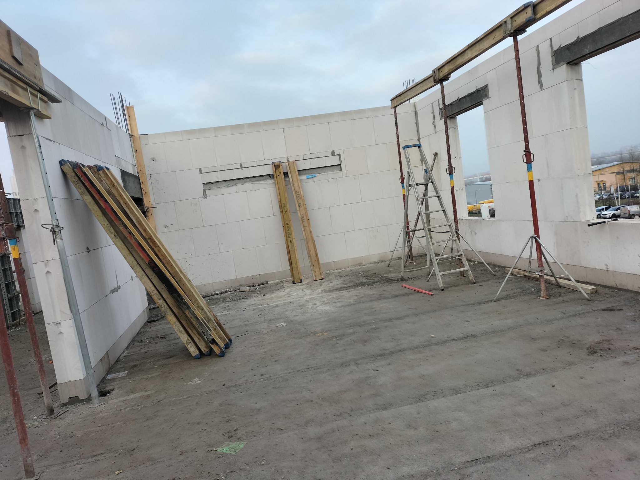 Wnętrze surowego domu z białych bloczków, widoczne drewniane stemple podtrzymujące strop, drabina i deski opierające się o ścianę, surowy stan deweloperski.