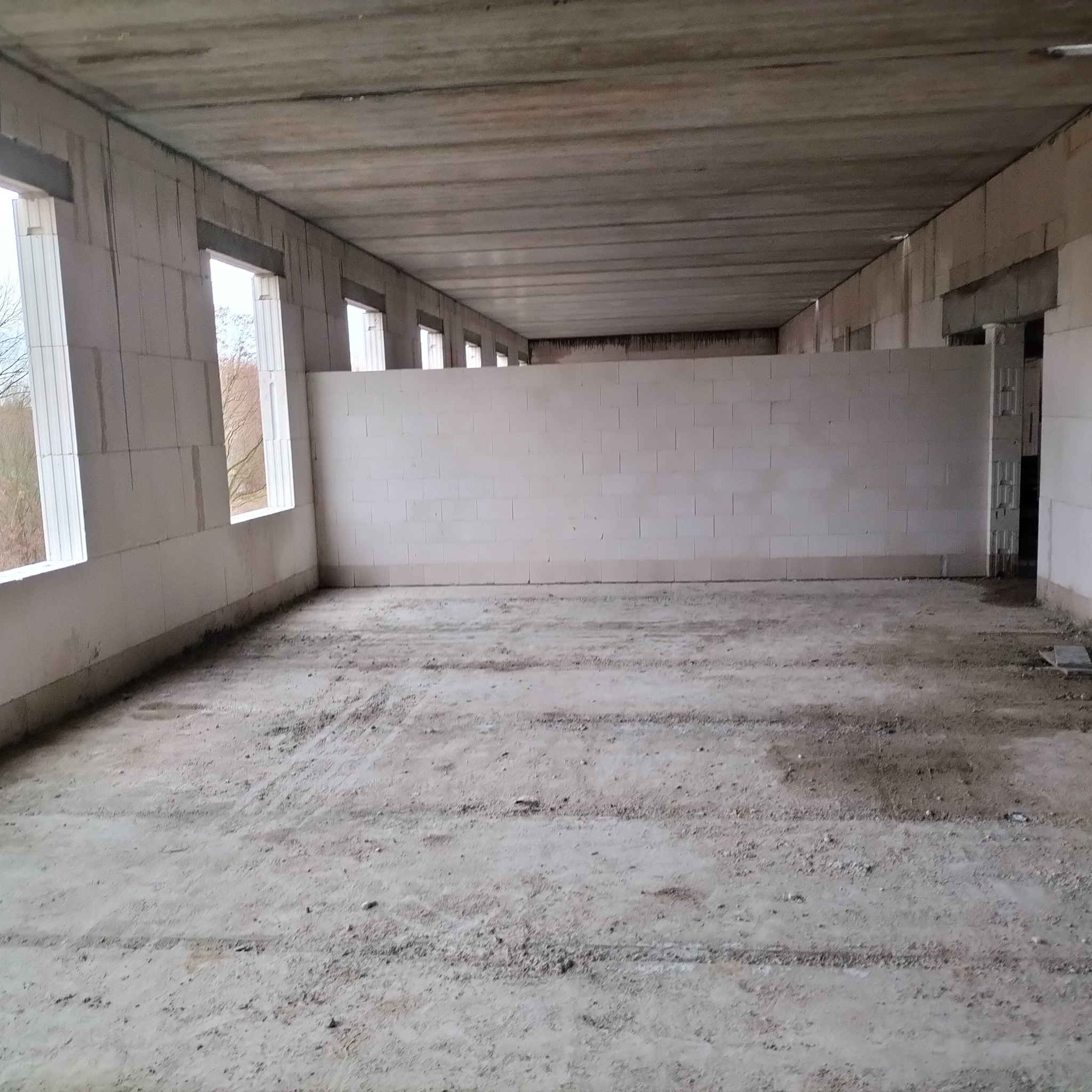 Wnętrze surowego budynku w trakcie budowy. Widoczne ściany z bloczków, okna i betonowa podłoga z resztkami materiałów budowlanych. Surowy stan deweloperski.