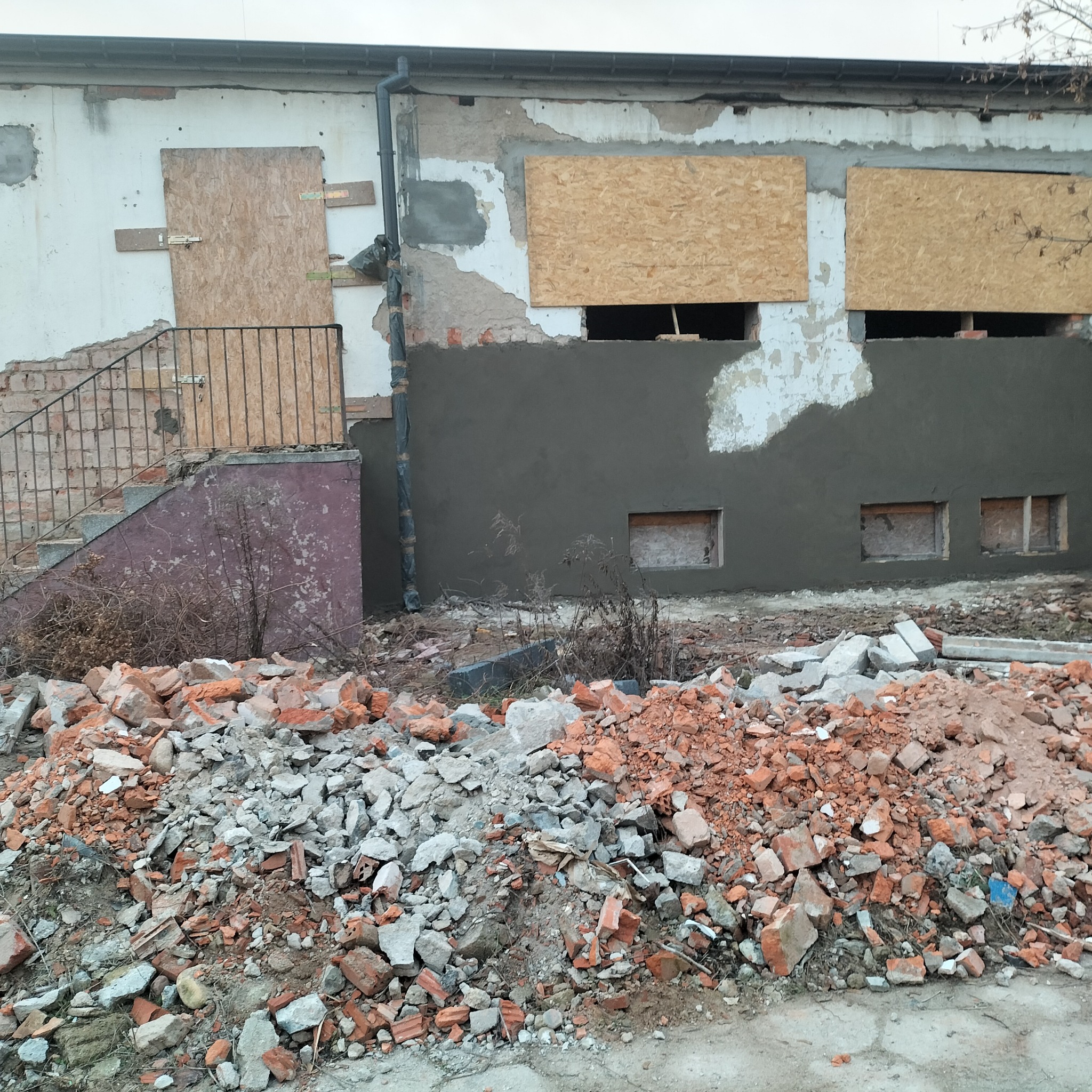Remont elewacji budynku z częściowo zakrytymi oknami płytami OSB i świeżo nałożoną warstwą tynku. Na pierwszym planie gruz budowlany.