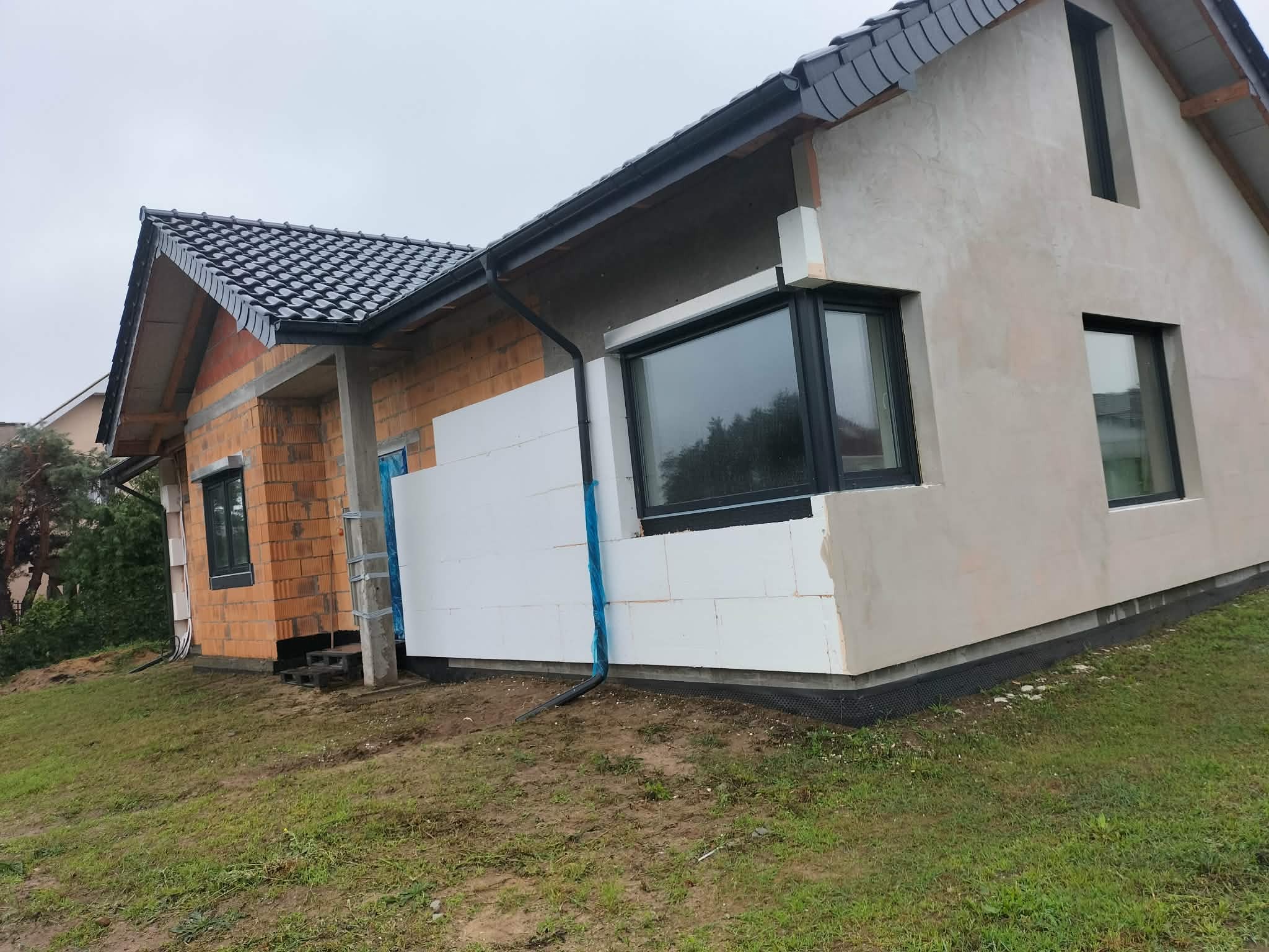 Dom w trakcie ocieplania: fragment ściany z cegły, fragment ocieplony styropianem, fragment otynkowany. Widoczne okna z czarnymi ramami i czarna rynna.