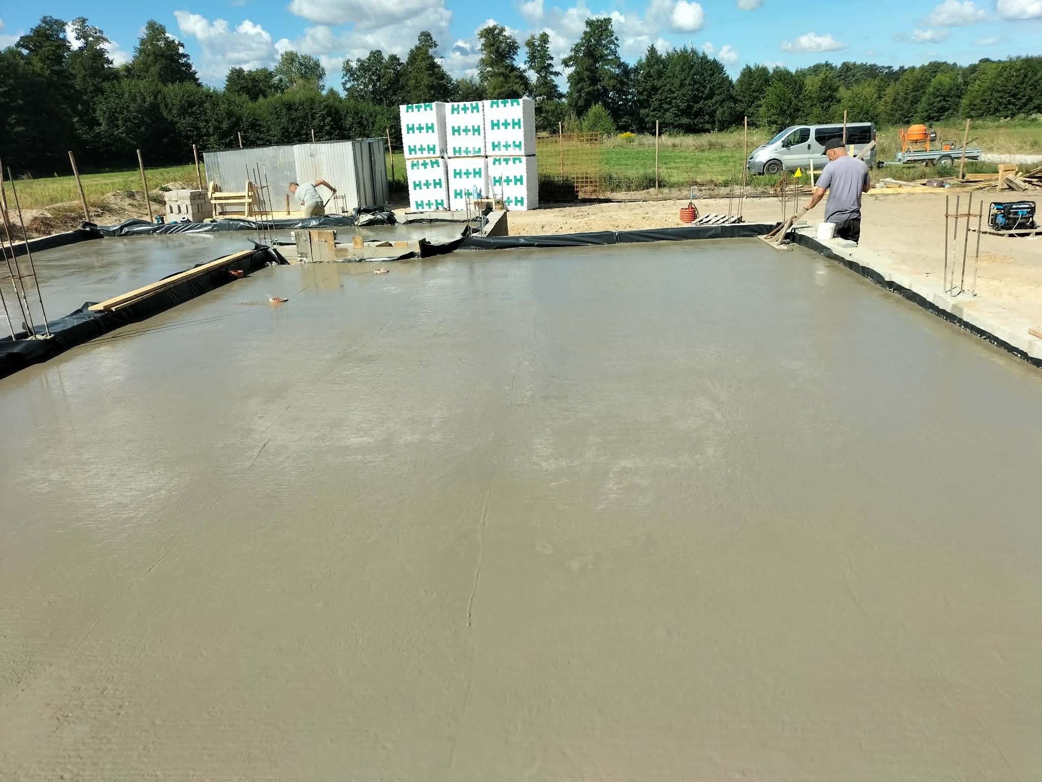 Wylewka fundamentowa w trakcie prac. Widoczny pracownik wyrównujący powierzchnię betonu, bloczki H+H, teren budowy i częściowo zaparkowany samochód dostawczy.