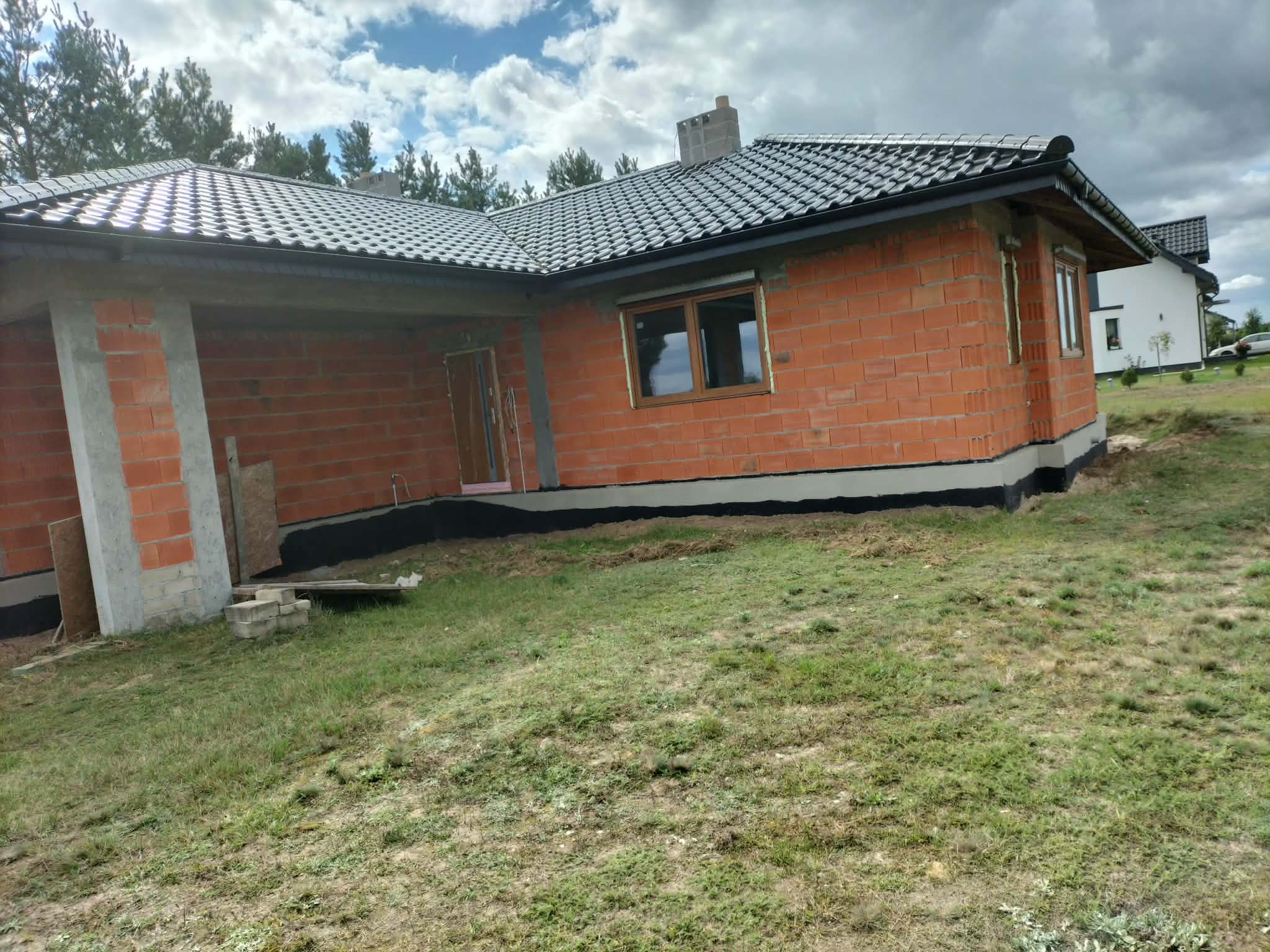 Surowy stan domu z czerwonej cegły z ciemną dachówką, widoczne okna z brązowymi ramami i fundament. Na pierwszym planie trawa i częściowo ułożone bloczki.