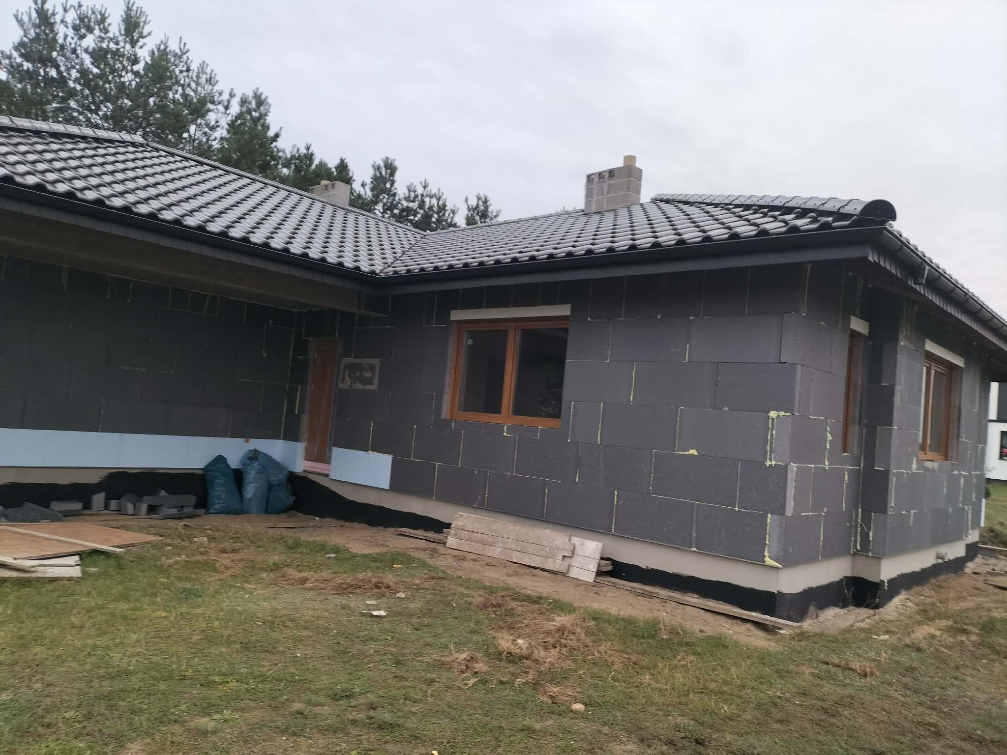 Budowa domu jednorodzinnego w trakcie ocieplania. Widoczne szare bloczki styropianowe na ścianach, okna z drewnianymi ramami i dach pokryty szarą dachówką.