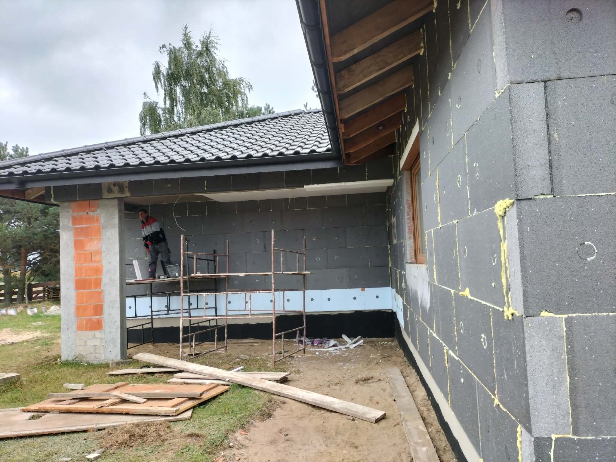 Budowa domu z szarego bloczka: widok na ścianę z oknem, częściowo ocieploną, z rusztowaniem i pracownikiem w tle. Widoczne deski i ceglany filar.