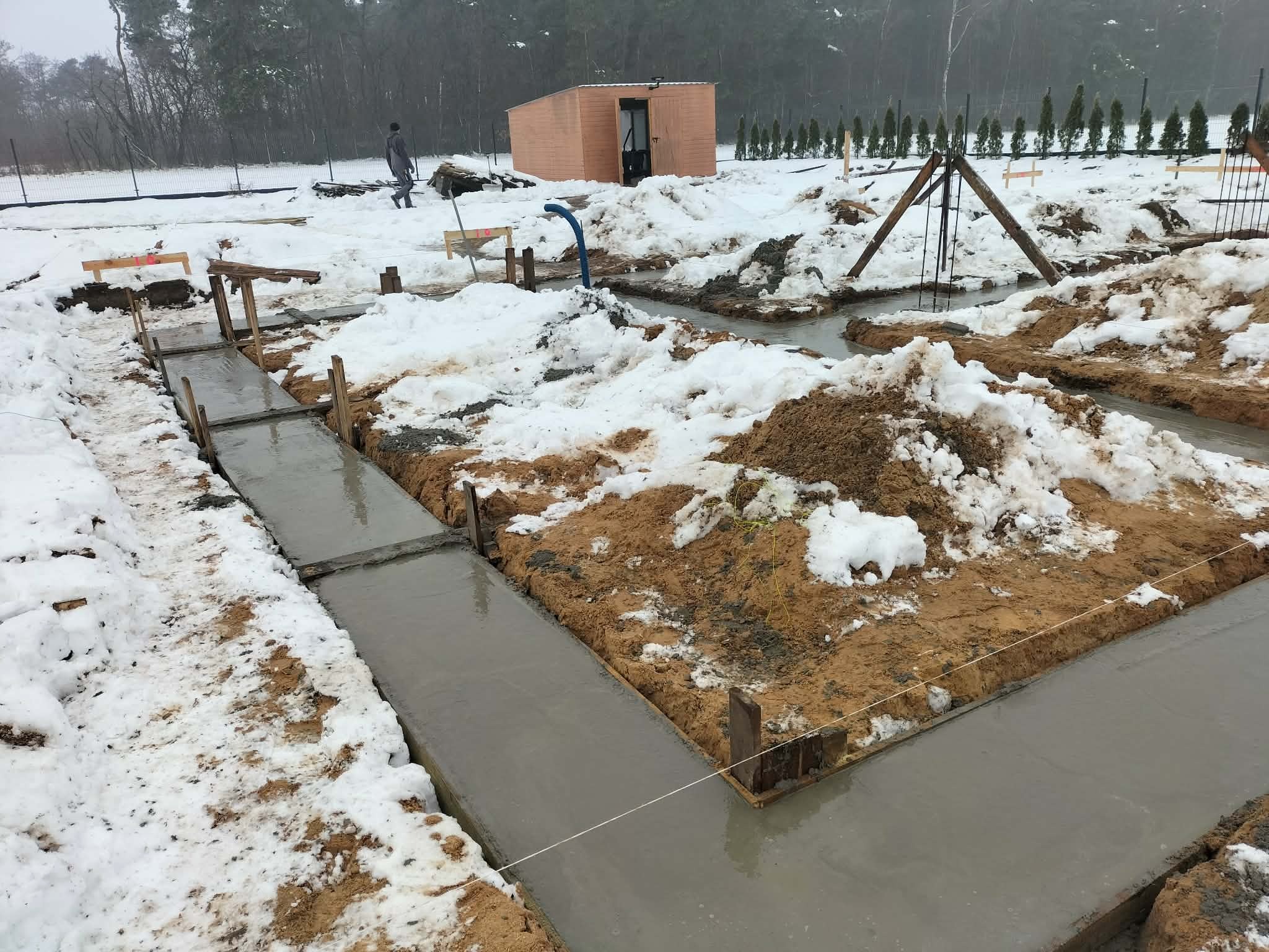 Zalane betonem ławy fundamentowe w zasypanym śniegiem wykopie. Widoczny szalunek i drewniane podpory. W tle barak budowlany i drzewa.