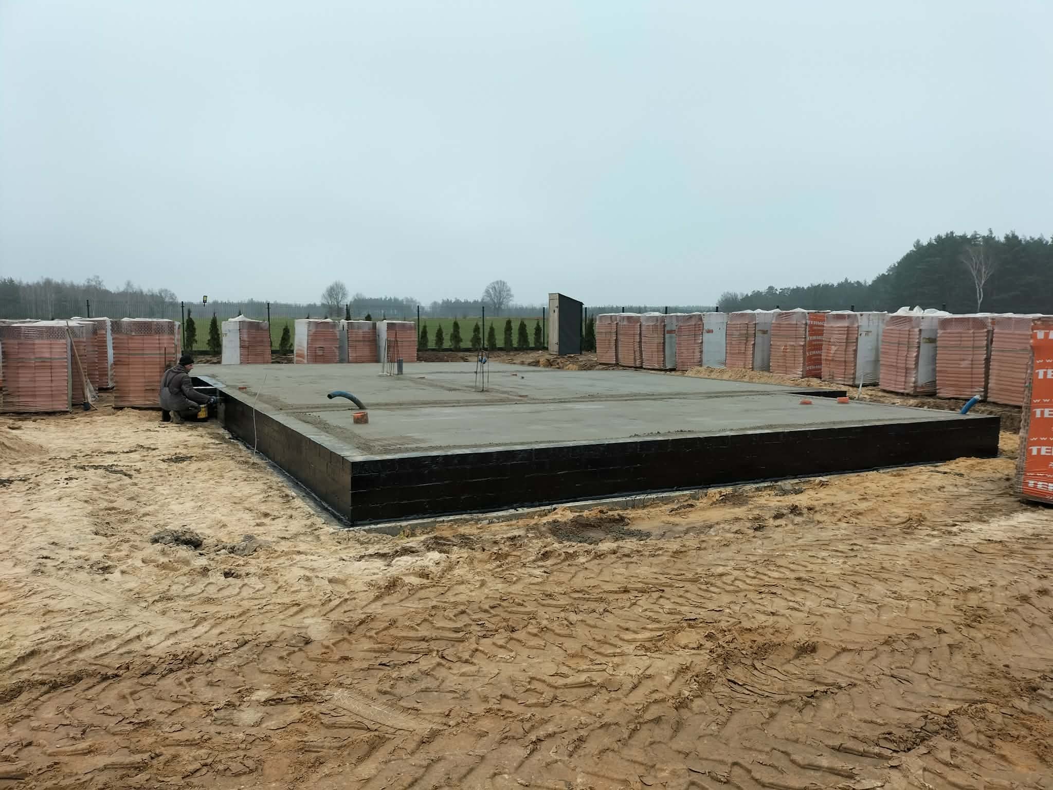 Zabezpieczone fundamenty domu jednorodzinnego, z betonową płytą i izolacją przeciwwilgociową, przygotowane do dalszych prac murarskich. W tle palety z cegłą.
