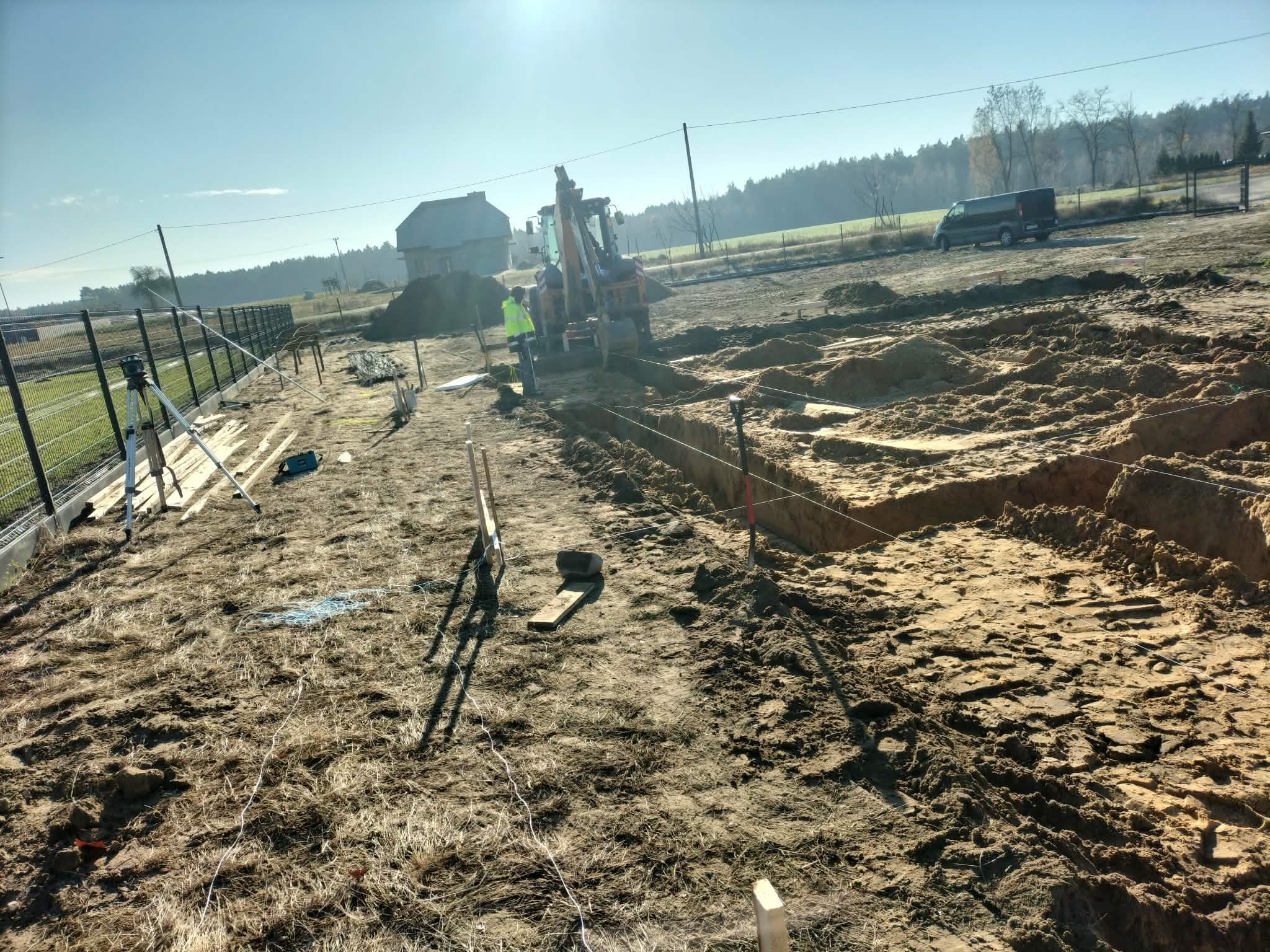 Wykop fundamentów pod budowę domu jednorodzinnego. W tle widoczny budynek, koparka i pracownik w odblaskowej kamizelce. Teren budowy ogrodzony siatką.