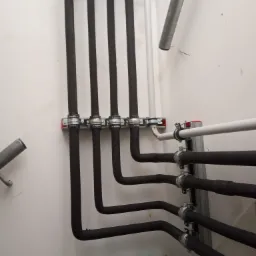 Czarny system rur grzewczych zamontowany na ścianie z użyciem metalowych uchwytów, widoczna butelka wody i miedziana rurka na podłodze.