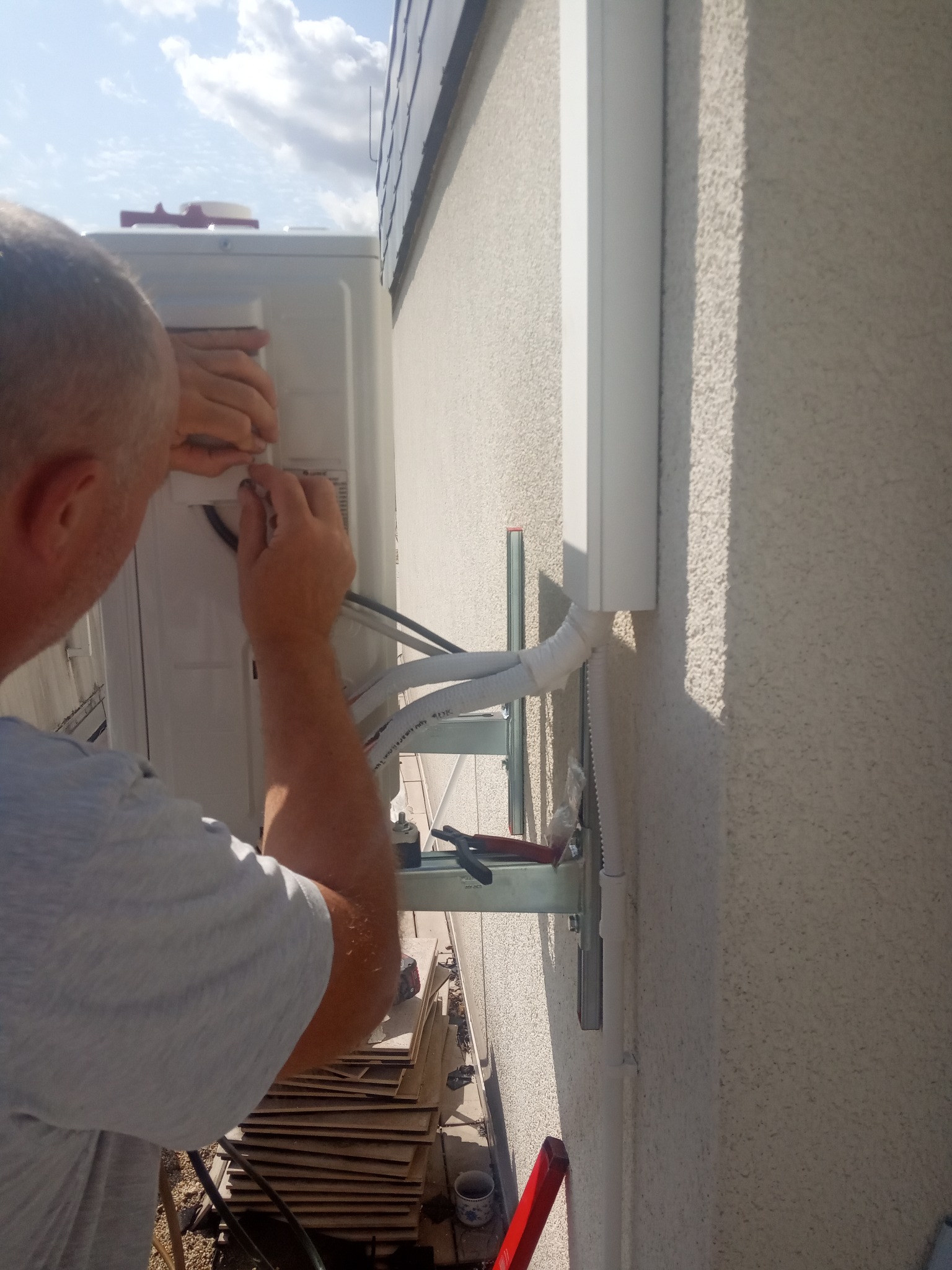 Elektryk podłącza przewody do zewnętrznej jednostki urządzenia chłodzącego, kable w białym kanale kablowym wraz z rurami freonowymi, montaż na ścianie budynku.