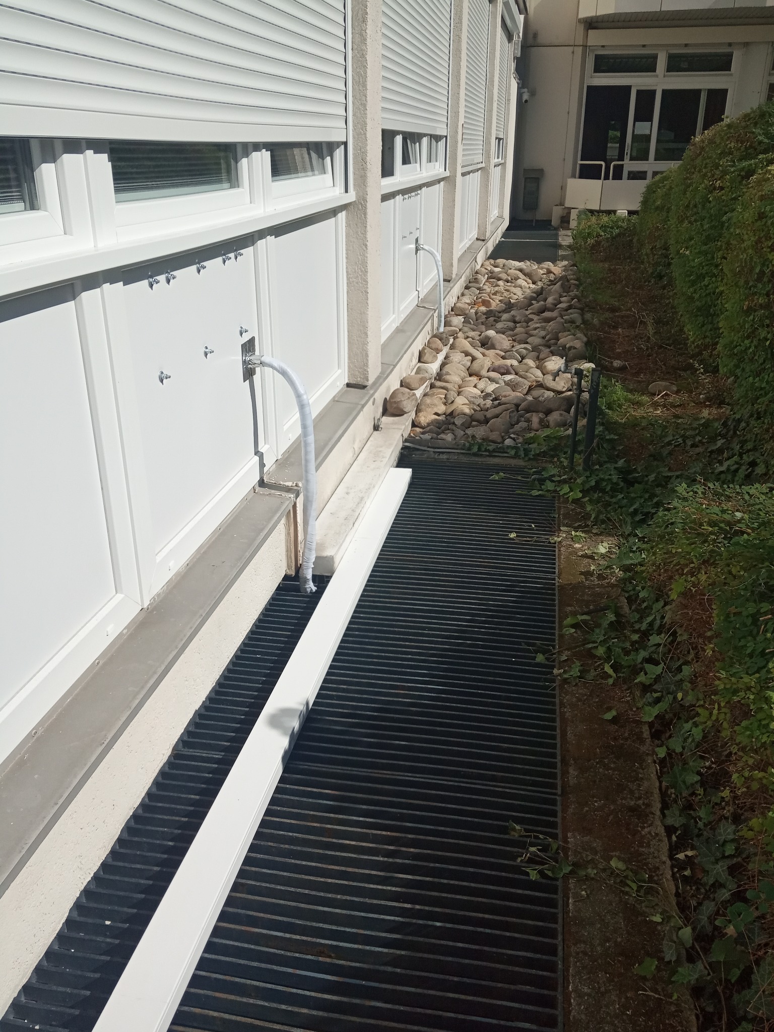 Wyjścia rur z urządzeń wewnętrznych poprowadzone z urządzeń przez ścianę do urządzenia zewnętrznego ukrytego pod kratką.