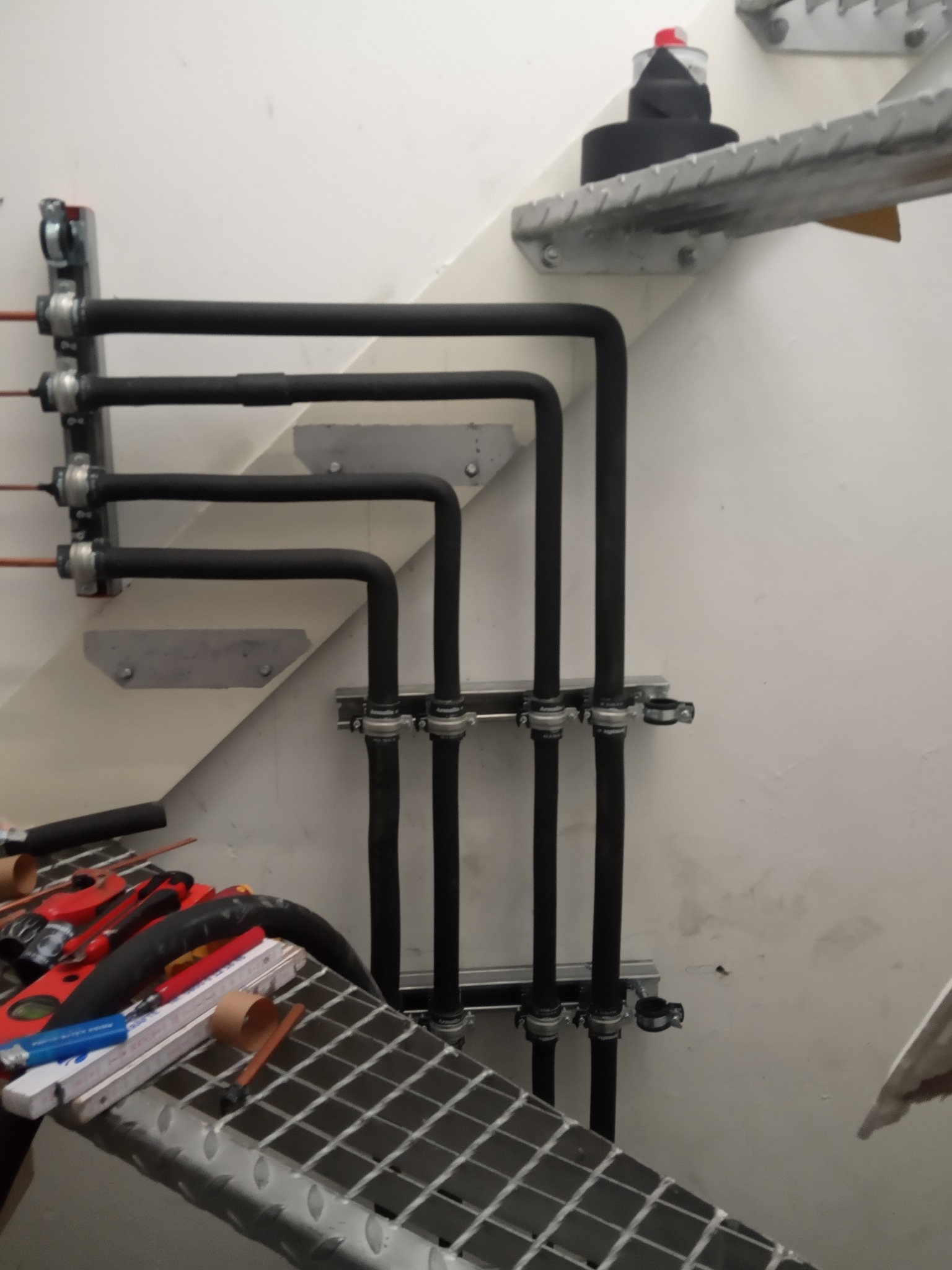 Cztery rury w izolacji, zamocowane do ściany na metalowych wspornikach, układają się równolegle wzdłuż schodów; widoczne narzędzia na metalowej platformie.