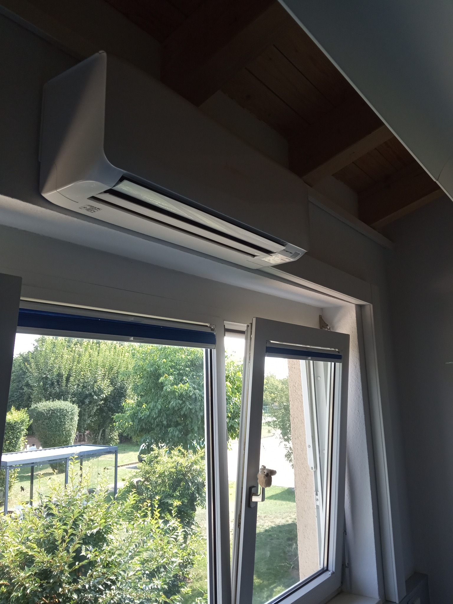 Biała jednostka wewnętrzna urządzenia zamontowana nad oknem z widokiem na zielony ogród z zewnątrz.