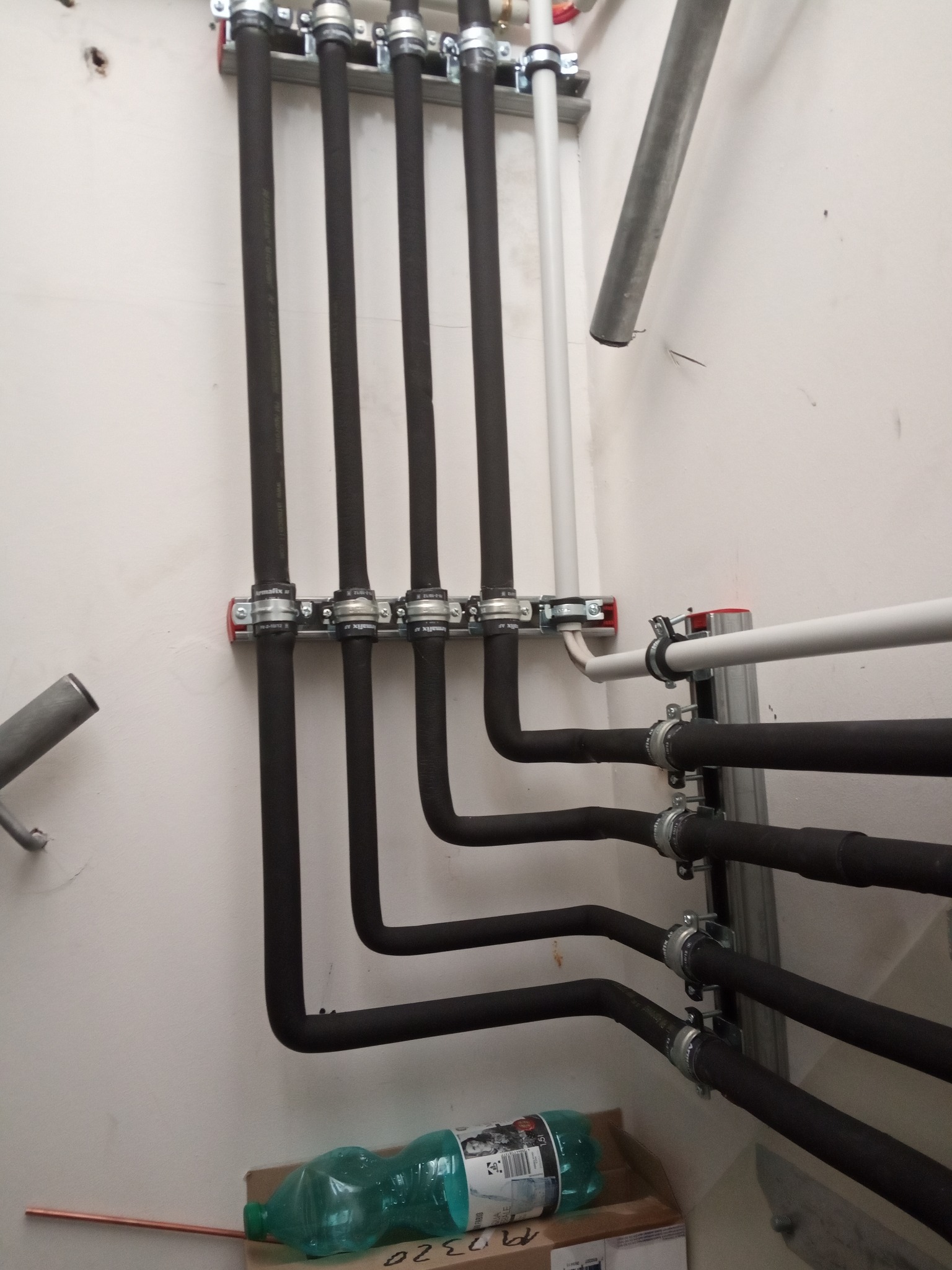 Czarny system rur grzewczych zamontowany na ścianie z użyciem metalowych uchwytów, widoczna butelka wody i miedziana rurka na podłodze.