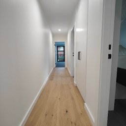 Piotr Smoliński - Jasny korytarz z drewnianą podłogą i świeżo pomalowanymi białymi ścianami, widok na drzwi wejściowe z zewnętrzną roślinnością w tle.