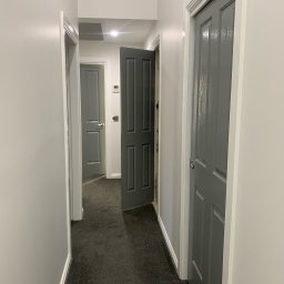 Piotr Smoliński - Korytarz po remoncie z szarymi ścianami i drzwiami, otwarte drzwi w głębi ukazują kolejne pomieszczenie, widoczna faktura świeżo pomalowanych ścian.