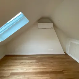 Pomalowane na biało poddasze ze skośnym oknem dachowym, klimatyzatorem i drewnianą podłogą w odcieniach brązu.