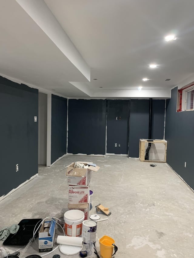 Pomieszczenie w trakcie malowania na ciemny kolor, widoczne niedomalowane fragmenty przy suficie i listwach, na podłodze puszki z farbą, wałki i inne narzędzia malarskie, surowy beton na podłodze.