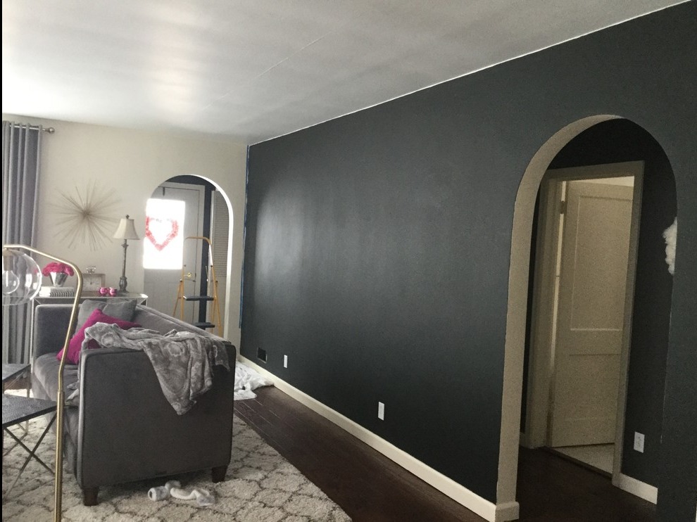 Świeżo pomalowana na grafitowo ściana w salonie z łukowatym przejściem, widoczny fragment sofy z poduszkami i kocykiem, drewniana podłoga i dywan w geometryczny wzór, drabina malarska w tle.