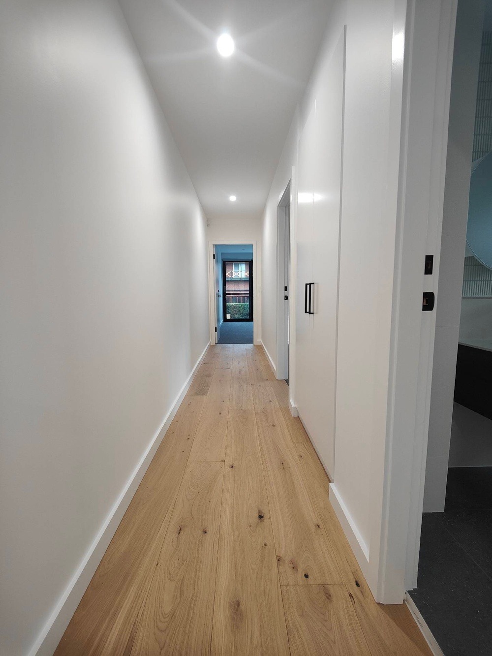 Jasny korytarz z drewnianą podłogą i świeżo pomalowanymi białymi ścianami, widok na drzwi wejściowe z zewnętrzną roślinnością w tle.