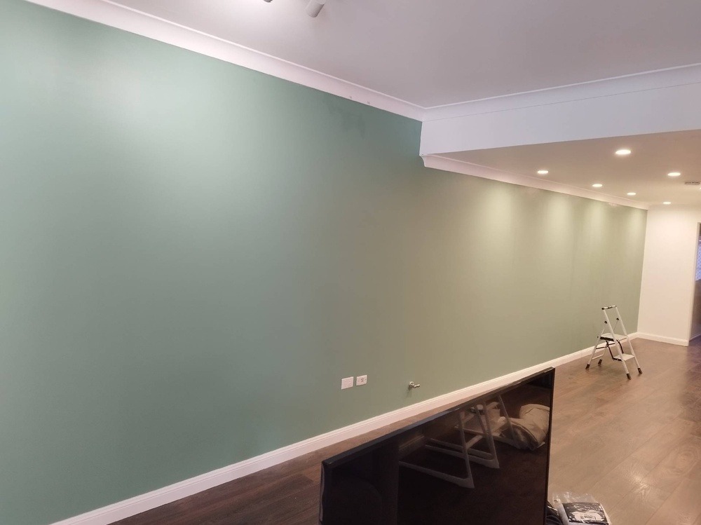 Świeżo pomalowana ściana w odcieniu zieleni z białymi listwami przypodłogowymi i sufitowymi, widoczna drabina malarska w tle, podłoga z paneli imitujących drewno.