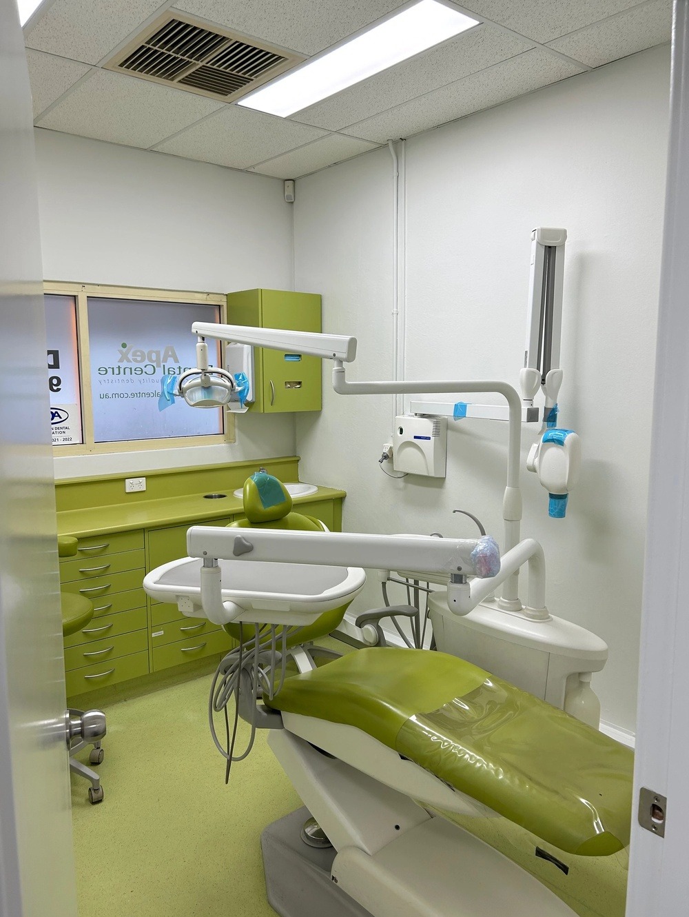 Jasne wnętrze gabinetu dentystycznego z zielonymi akcentami, widoczny fotel dentystyczny z osprzętem, szafki i umywalka w odcieniach zieleni, białe ściany i sufit.